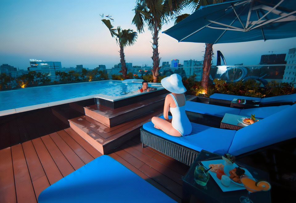 a woman in a white hat is sitting on lounge chairs near a pool , overlooking the city at Alagon Saigon Hotel & Spa