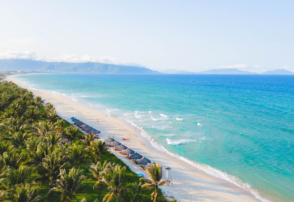 a beautiful beach with clear blue water and white sand , surrounded by palm trees and mountains at Movenpick Resort Cam Ranh