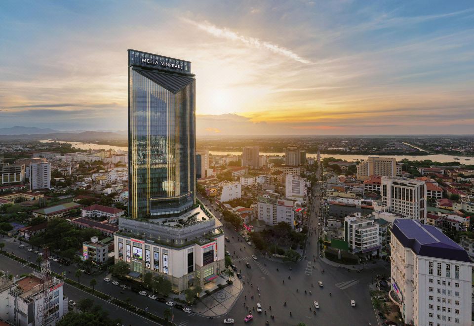 a large building with a unique design is surrounded by smaller buildings and trees , with the sun setting in the background at Melia Vinpearl Hue