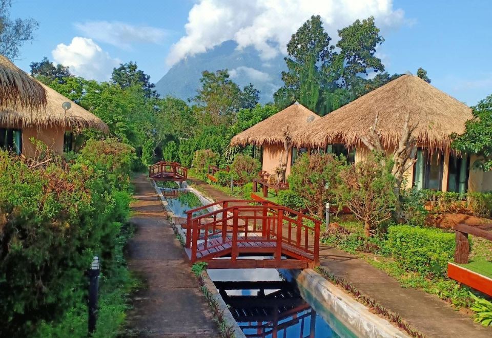 a serene garden with a red bridge and several thatched - roof huts in the background , set against a backdrop of mountains at Aurora Resort Chiangdao