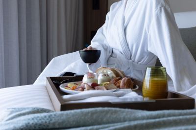 a person in a robe is sitting at a bed with a tray of food and a glass of wine at Oval Hotel at Adelaide Oval, an EVT hotel