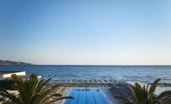 a swimming pool surrounded by palm trees , with the ocean visible in the background , under a clear blue sky at Civitel Creta Beach