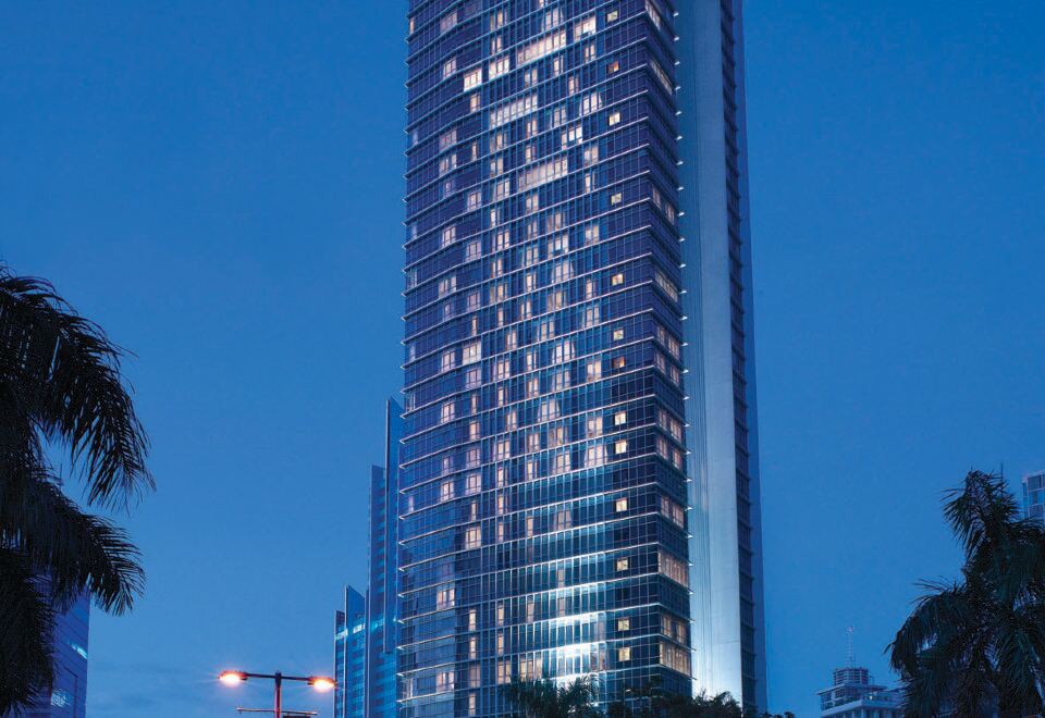 a tall , modern building with many windows is lit up at night , surrounded by palm trees at Oakwood Premier Cozmo Jakarta