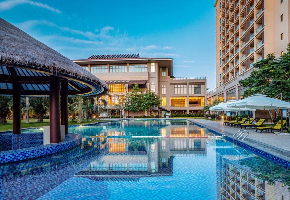 a modern hotel with a large swimming pool and umbrellas in front of it , surrounded by lush greenery at Sanya Xizang Hotel