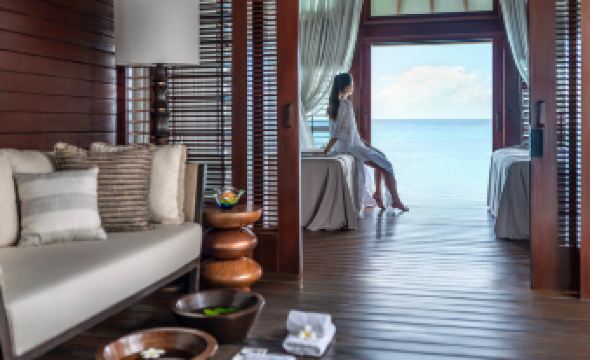 a woman in a white dress sitting on a couch with a view of the ocean , while wearing a mask at Four Seasons Resort Maldives at Kuda Huraa