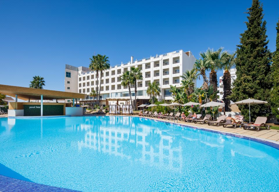 a large outdoor swimming pool surrounded by palm trees , with a hotel in the background at AP Maria Nova Lounge