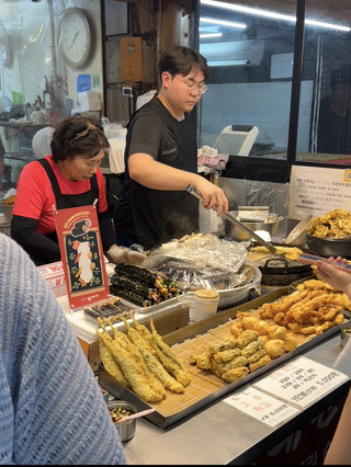 第一次走進首爾傳統市集,就能感受到和大型百貨商場完全不同的氛圍。入口兩旁擺滿新鮮蔬果、韓式小菜、乾貨、海鮮和各式熟食,攤販熱情招呼客人,空氣中瀰漫著現烤魚、辣炒年糕、糖餅與麻油香氣,光是一路慢慢逛,就讓人忍不住一直停下腳步。

我最喜歡的是市集保留了濃厚的在地生活感,不只是觀光客會來,更多的是附近居民每天採買食材、吃早餐或午餐的地方。看著老闆熟練地切肉、現做煎餅、包紫菜飯捲,能感受到這裡幾十年累積下來的人情味,也比熱門商圈更能體驗真正的韓國日常。

市集裡的美食選擇非常多,而且價格普遍比觀光區親民。這次品嚐了辣炒年糕、魚板湯、韓式煎餅、血腸、糖餅以及現炸熱狗,每一攤都有自己的特色。尤其剛起鍋的綠豆煎餅外酥內軟,搭配現磨豆漿或米酒,更能感受到韓國傳統飲食文化。很多攤位都是現點現做,即使需要稍微等待,也很值得。

除了美食之外,市集還販售韓國傳統點心、泡菜、海苔、人參、韓式餐具、生活用品、服飾及季節水果。如果不趕時間,很推薦慢慢逛,常常會發現不少意外驚喜。有些攤販還會提供試吃,不用急著購買,多比較幾家再決定,通常都能找到品質不錯又價格合理的商品。

讓我印象最深刻的是攤販與客人的互動,即使語言不完全相通,老闆仍會用簡單英文、手勢或翻譯軟體耐心介紹商品,有時還會多送一點小菜或請客人試吃,整個購物過程十分輕鬆,也讓人感受到韓國市場特有的人情味。

如果是第一次造訪首爾,我很推薦安排半天到一天走訪傳統市集,而不是只逛明洞、弘大或聖水。每個市集都有自己的特色,有些以小吃聞名,有些以海鮮、生鮮或服飾著稱,即使是同樣的韓式料理,不同市集也能吃到不同風味。

旅遊小建議:

· 建議避開中午尖峰時段,人潮較少,逛起來更舒服。
· 攜帶一些現金,部分攤販雖可刷卡,但現金仍較方便。
· 空著肚子前往,可以一次品嚐更多韓國在地小吃。
· 若想購買泡菜、海苔或伴手禮,建議多比較幾家價格與份量。
· 市集通道較窄,假日人潮很多,建議穿著舒適好走的鞋子。

整體而言,首爾傳統市集不只是購物或吃美食的地方,更是一個能深入了解韓國文化與居民生活的空間。沒有過度商業化的包裝,取而代之的是熱鬧叫賣聲、現做料理的香氣,以及充滿溫度的人情交流。這趟市集之旅讓我留下非常深刻的印象,也成為整趟首爾旅行中最有生活感、最值得回味的一段回憶。每次回想起來,腦海中浮現的不是華麗景點,而是那些充滿煙火氣息的小攤、美味的街頭料理,以及市集裡最真實的首爾日常。