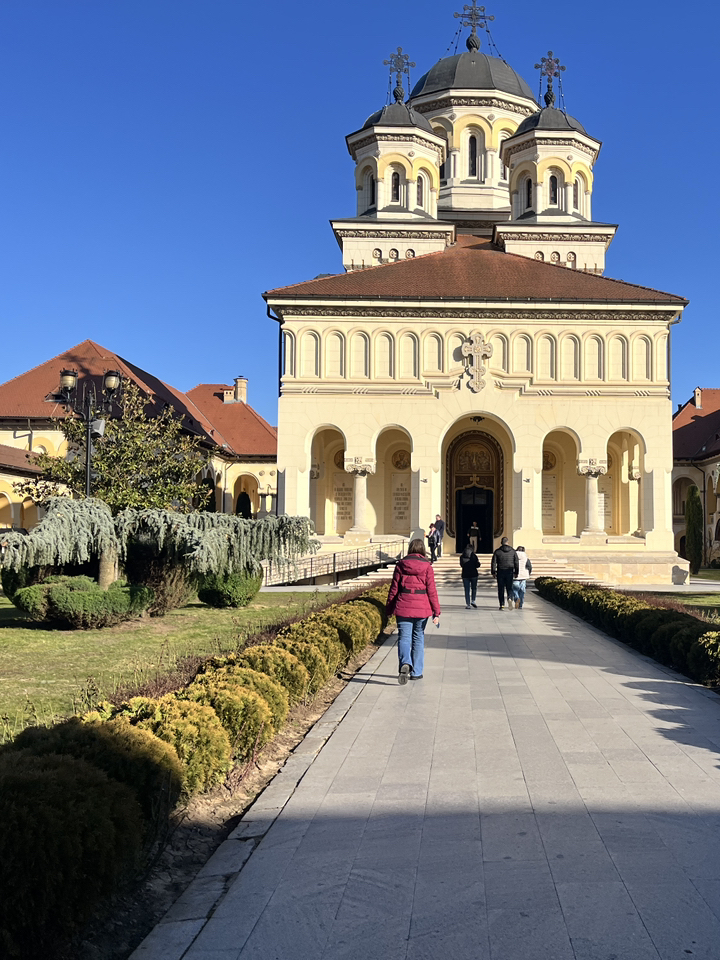 Memasuki Benteng Alba Carolina lebih seperti berjalan melalui lingkaran waktu di mana tiga era berbeda hidup berdampingan, daripada sekadar mengunjungi museum. Terletak di jantung Transylvania, benteng berbentuk bintang berujung tujuh ini adalah yang terbesar di Rumania, sebuah mahakarya bergaya Vauban yang telah menjaga rahasia negara selama berabad-abad.
Perjalanan Anda dimulai di Gerbang Ketiga, yang paling megah dari enam pintu masuk Barok. Dihiasi dengan relief rumit dan patung-patung tokoh mitologi, gerbang ini berfungsi sebagai portal besar menuju taman bermain bersejarah seluas 140 hektar. Saat Anda menyusuri jalan setapak berbatu pada tahun 2026, Anda akan menyaksikan Pergantian Penjaga pada siang hari. Para prajurit dengan seragam Austria abad ke-18 berbaris dengan presisi ritmis, senapan dan genderang mereka bergema di dinding bata tebal—sebuah tontonan teatrikal yang membuat era Habsburg terasa sangat nyata.

Cakrawala benteng didominasi oleh dua bangunan raksasa yang berdiri berdampingan. Katedral St. Michael, sebuah bangunan Katolik Roma berusia lebih dari 800 tahun, menyimpan makam John Hunyadi dan memiliki menara tertinggi di wilayah tersebut. Hanya sepelemparan batu dari sana terdapat Katedral Penobatan, sebuah bangunan neo-Bizantium yang indah tempat Raja Ferdinand dan Ratu Marie dinobatkan pada tahun 1922, menandai kelahiran simbolis Rumania Raya.
Bagi mereka yang berkunjung pada Juli 2026, jangan lewatkan festival Barockada, di mana musik kuno memenuhi udara, berpadu sempurna dengan suasana megah benteng. Jika Anda lebih menyukai suasana yang lebih tenang, Jalur Tiga Benteng memungkinkan Anda untuk melihat reruntuhan castrum Romawi dan tembok abad pertengahan yang mendahului struktur saat ini.
Baik Anda sedang menikmati kopi di dekat Balai Persatuan—tempat penandatanganan penyatuan tahun 1918—atau bersepeda di sepanjang parit, Alba Iulia menawarkan perpaduan langka antara kesungguhan dan pesona. Ini adalah tempat di mana sejarah tidak hanya diingat; Itu sudah terjadi.

