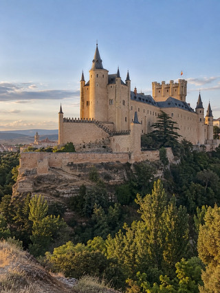 Segovia Is the Perfect Day Trip from Madrid — But It Deserves More Than a Few Hours 🇪🇸🏰

Segovia feels almost designed for a perfect Spanish day trip: a monumental Roman aqueduct in the middle of the city, medieval streets, a huge Gothic cathedral, an extraordinary castle and restaurants serving one of Castilla y León’s most famous dishes.

The historic city and its Roman Aqueduct are UNESCO World Heritage, and almost everything visitors want to see can be explored on foot.

It is very easy to visit from Madrid, but my biggest recommendation is not to rush back immediately after seeing the Aqueduct and Alcázar. Segovia becomes particularly beautiful when the day-trippers begin leaving.

ℹ️ Practical Visit Information

📍 Location: Castilla y León, Spain
🚆 From Madrid: High-speed trains make Segovia an easy day trip
🗓️ Recommended stay: 1 full day; overnight if you want a quieter experience
🏛️ Don’t miss: Roman Aqueduct, Cathedral, Jewish Quarter and Alcázar
👟 Getting around: Walking is the best option
⛰️ Terrain: Expect hills, stairs and cobbled streets
🍽️ Must try: Cochinillo asado and ponche segoviano
📸 Best photography: Postigo del Consuelo, Mirador de la Pradera de San Marcos and Plaza Mayor
🌅 Best time: Morning for monuments and late afternoon for viewpoints
💡 Best tip: Walk from the Aqueduct to the Alcázar instead of jumping between attractions by transport
⚠️ Important: Reserve the Alcázar in advance during busy weekends and holidays.

🚆 Getting to Segovia from Madrid

The fastest option is the high-speed train from Madrid to Segovia-Guiomar.

One important detail: Segovia-Guiomar station is outside the historic centre.

Don’t leave the station expecting the Roman Aqueduct to be around the corner. You’ll need a bus or taxi to reach central Segovia.

If you prefer arriving directly into the urban area, intercity buses from Madrid are another practical option.

For a day trip, leave Madrid early and give yourself the entire day.

🏛️ Start at the Roman Aqueduct

Nothing really prepares you for turning a corner and finding an enormous Roman aqueduct crossing the city.

The Aqueduct has 167 arches, and its massive granite blocks were assembled without mortar.

Start around Plaza del Azoguejo, but don’t photograph it only from ground level.

Climb the stairs towards the Postigo del Consuelo.

The official Spain tourism guide specifically recommends this elevated position for one of the best views of the monument.

📸 The Best Aqueduct Photo Isn’t from Directly Underneath

From Plaza del Azoguejo, everyone photographs upwards.

Instead, climb beside the Aqueduct.

From above, you can capture the arches stretching across Segovia with rooftops and surrounding landscape behind them.

Go early if you want cleaner photographs.

Then return after dark.

The illuminated Aqueduct against the night sky is completely different.

🚶 Follow Calle Real

One of Segovia’s best walks requires almost no planning.

Follow the traditional route from the Aqueduct towards the Alcázar through the historic centre.

Along the way you’ll pass Casa de los Picos, Plaza de Medina del Campo, San Martín, the Cathedral and eventually the Alcázar. Segovia’s tourism authority specifically promotes this historic axis as the route «Del Acueducto al Alcázar».

Don’t hurry.

Look at the façades and decorative patterns on the buildings as you walk.

⛪ Stop at Segovia Cathedral

The Cathedral dominates Plaza Mayor and is sometimes nicknamed the «Lady of the Cathedrals».

Even if you’re not normally interested in churches, go inside.

The scale, Gothic architecture and stained glass make it worthwhile.

Then spend some time around Plaza Mayor.

This is a good place for coffee before continuing towards the Alcázar.

✡️ Walk Through the Jewish Quarter

The former Jewish Quarter is very close to the Cathedral but easy to overlook.

Explore Calle de la Judería Vieja and the surrounding narrow streets.

You can also see the former Main Synagogue and, farther away, the area associated with Segovia’s historic Jewish cemetery.

It provides an important additional layer to the city’s medieval history.

🏰 The Alcázar Looks Like a Fairytale Castle

The Alcázar of Segovia occupies a spectacular rocky position above the surrounding valleys.

Its pointed towers, slate roofs and ship-like profile make it one of Spain’s most distinctive castles.

Explore the royal rooms, armoury and chapel.

But if you’re physically comfortable with stairs, add the Tower of Juan II.

The official tourism route notes a climb of 152 steps and spectacular views towards Vera Cruz, El Parral and the surrounding mountains.

The staircase is narrow, so don’t underestimate it.

🌳 Find the Best View of the Alcázar

Here’s the photograph I wouldn’t miss.

After visiting the castle, descend towards the Pradera de San Marcos.

From below, the Alcázar rises above the trees and rocky cliff like the castle from a storybook.

Late afternoon is particularly beautiful.

This perspective also shows why the castle looks so different from inside the historic centre.

🍽️ Try Cochinillo Asado

Segovia is famous for cochinillo asado — roast suckling pig.

Traditionally, the skin should be extremely crisp while the meat remains tender.

It’s rich, so arrive hungry.

Another local classic is lechazo, roast milk-fed lamb. Spain’s official tourism guide recommends both among Segovia’s traditional specialities.

If you’re travelling with someone, consider sharing dishes rather than ordering an enormous meal immediately before climbing the Alcázar tower.

🍰 Save Room for Ponche Segoviano

Don’t leave after the cochinillo.

Try ponche segoviano, the city’s traditional dessert.

It’s a rich layered sweet typically involving sponge cake, cream and marzipan.

Order a portion with coffee rather than buying a huge one unless you have a serious sweet tooth.

It’s the perfect excuse to stop at a traditional pastry shop during the afternoon.

🌿 Walk Down to the Eresma

If you have a full day, go beyond the standard Aqueduct–Cathedral–Alcázar itinerary.

Descend towards the Eresma River.

You can explore the green belt around the historic city and see places such as the Church of Vera Cruz, Monastery of El Parral and Fuencisla area. Spain’s official tourism itinerary recommends this lower route for its scenery and alternative views of Segovia.

You’ll also encounter far fewer visitors.

📸 Best Photography Spots

For the Aqueduct: Postigo del Consuelo.

For the Alcázar: Pradera de San Marcos.

For rooftops and mountains: climb the Tower of Juan II.

For medieval streets: explore the Jewish Quarter early.

For the Cathedral: photograph from Plaza Mayor and surrounding streets.

And stay until evening if possible.

The Aqueduct illuminated after sunset is a fantastic final photograph.

💡 Tips That Will Make Your Visit Better

Take an early train from Madrid.

Wear comfortable shoes because Segovia has constant changes in elevation — something Spain’s official tourism guide also warns visitors about.

Walk from the Aqueduct to the Alcázar.

Book major monuments ahead during busy periods.

Have lunch slightly earlier or later than peak time if you haven’t reserved.

Go down to Pradera de San Marcos.

Try both cochinillo and ponche segoviano.

Carry water during summer.

And don’t schedule your return to Madrid too early.

⚠️ Things to Avoid

Don’t assume Segovia-Guiomar station is in the historic centre.

Don’t visit only the Aqueduct.

Don’t wear uncomfortable shoes.

Don’t eat a huge cochinillo lunch immediately before climbing 152 stairs.

Don’t spend the entire day following large tour groups.

Don’t climb onto protected monuments for photographs.

Don’t skip the Jewish Quarter.

And don’t rush through the Alcázar simply because you have a train back to Madrid.

🌤️ Best Time to Visit

Spring and autumn are excellent for walking.

Summer provides long days but can become hot around midday.

Winter is colder — Segovia sits at roughly 1,000 metres above sea level — but the historic streets can feel particularly atmospheric.

Weekdays are preferable if you want fewer visitors.

Whatever the season, check the weather before leaving Madrid. Conditions can feel noticeably different.

❤️ Final Thoughts

Segovia is one of the easiest places to recommend from Madrid because the amount of history packed into such a walkable city is remarkable.

Start beneath a 2,000-year-old Roman Aqueduct, walk through medieval streets and the Jewish Quarter, eat cochinillo, visit one of Spain’s most distinctive castles and finish the afternoon looking back at the Alcázar from the valley.

You can technically see Segovia in a few hours.

But give it a full day and you’ll discover that the Aqueduct is only the beginning. 🇪🇸🏛️🏰 #travel #travel2026 #segovia 