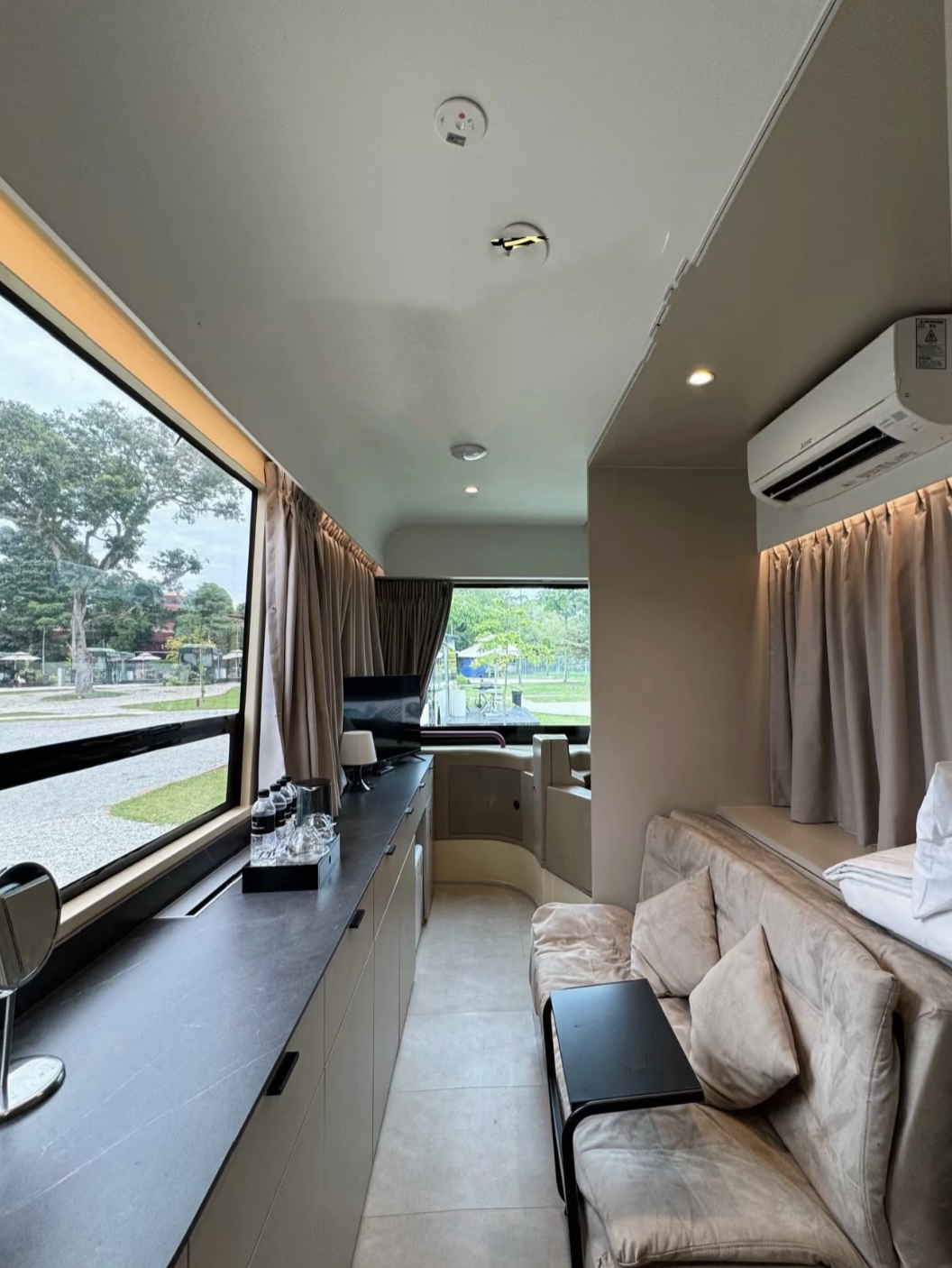 Terletak di Changi Village yang rindang, The Bus Collective adalah resor pertama di Asia Tenggara yang dibangun di dalam bus umum SBS yang sudah pensiun. Setiap bus telah diubah menjadi suite seluas 45 meter persegi, lengkap dengan meja marmer, pancuran hujan, bathtub, tempat tidur mewah, serta kursi pengemudi dan setir asli yang tetap dipertahankan—nostalgia bertemu kenyamanan di atas roda.

Saya menginap di salah satu tata letak ramah keluarga yang diparkir di tepi sungai. Ruangannya terasa rapi dan praktis, dengan sofa bed yang nyaman, TV pintar, Wi-Fi yang kuat (meskipun kadang-kadang dilaporkan terputus), dan dapur kecil yang berguna. Kursi pengemudi menjadi favorit yang menyenangkan—cocok untuk anak-anak—atau untuk mengenang perjalanan bus masa kecil.

Teras luar adalah sorotan utama—dilengkapi dengan tempat panggangan, meja makan, payung, dan pemandangan pesawat yang mendarat di dekatnya. Malam-malam yang dihabiskan untuk memanggang sate dan makanan laut terasa sangat santai. Jika Anda lebih suka makanan lokal, Pusat Jajanan Changi Village yang terkenal hanya berjarak dua menit berjalan kaki melalui pintu gerbang.

Sebagian besar tamu menyukai suasananya: tenang, ramah keluarga, dan menyenangkan. Ulasan memuji keunikannya, staf yang ramah, kamar yang bersih, dan malam BBQ yang berkesan di bawah langit. Skor berkisar antara 8–9/10—pengalaman staycation yang unik dekat dengan alam dan bandara.

Jika Anda mencari staycation yang merupakan perpaduan antara petualangan unik dan kenyamanan, The Bus Collective memberikan pengalaman tersebut. Ini bukan kemewahan ultra, dan ruangnya cukup sempit—tetapi ini menyenangkan, berkesan, dan ideal untuk keluarga atau siapa saja yang pernah bermimpi tidur di dalam bus. Hanya saja, jangan datang dengan harapan mendapatkan suite sebesar kondominium—atau keheningan tanpa suara.