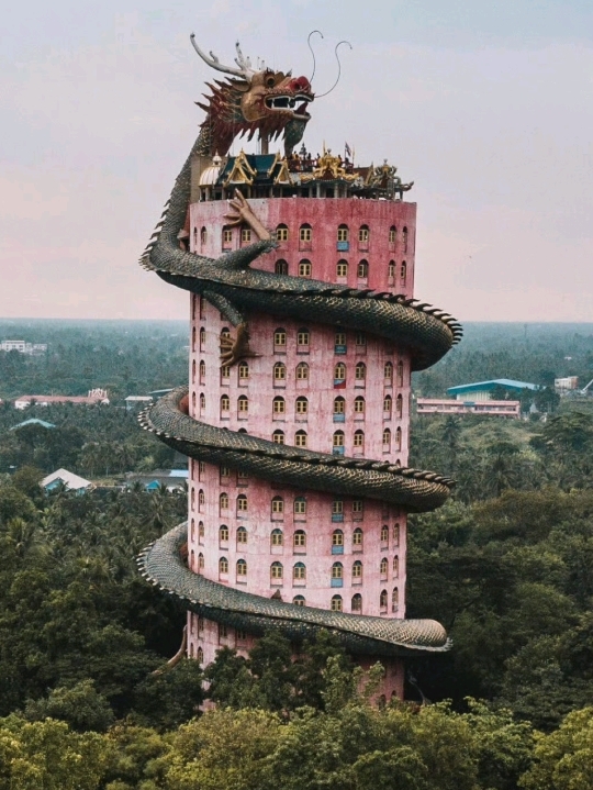 In the outskirts of Bangkok, Wat Sam Phran rises like something from a dream that didn’t bother checking reality first.

A massive pink cylindrical tower stands in the forest, wrapped by a giant green dragon that coils from ground to sky as if the entire structure is its resting form rather than its enclosure. The scale feels theatrical, almost cinematic, but also strangely quiet because it sits away from the dense urban rhythm of Bangkok.

Unlike traditional Thai temples that spread horizontally across courtyards and gilded halls, this one pulls the eye upward in a single continuous spiral. Inside the dragon’s body, a stairway runs through the structure, turning the climb into a passage through scales, shadow, and filtered light.

From a distance, the temple feels almost unreal—like a digital rendering placed into a real forest. Up close, the texture of paint, concrete, and carved detail makes it unexpectedly physical, even slightly weathered, as if myth had been built and then left to age in tropical humidity.

📍 Location: Wat Sam Phran
🏙️ Nearby city: Bangkok
🐉 Signature feature: giant dragon wrapping a pink cylindrical tower
🌿 Setting: suburban forest edge, quiet and isolated
📸 Best moment: late afternoon light when the dragon silhouette becomes more pronounced

Some temples are built to be worshipped.

Wat Sam Phran feels like it was built to be remembered. 🐉🌸✨