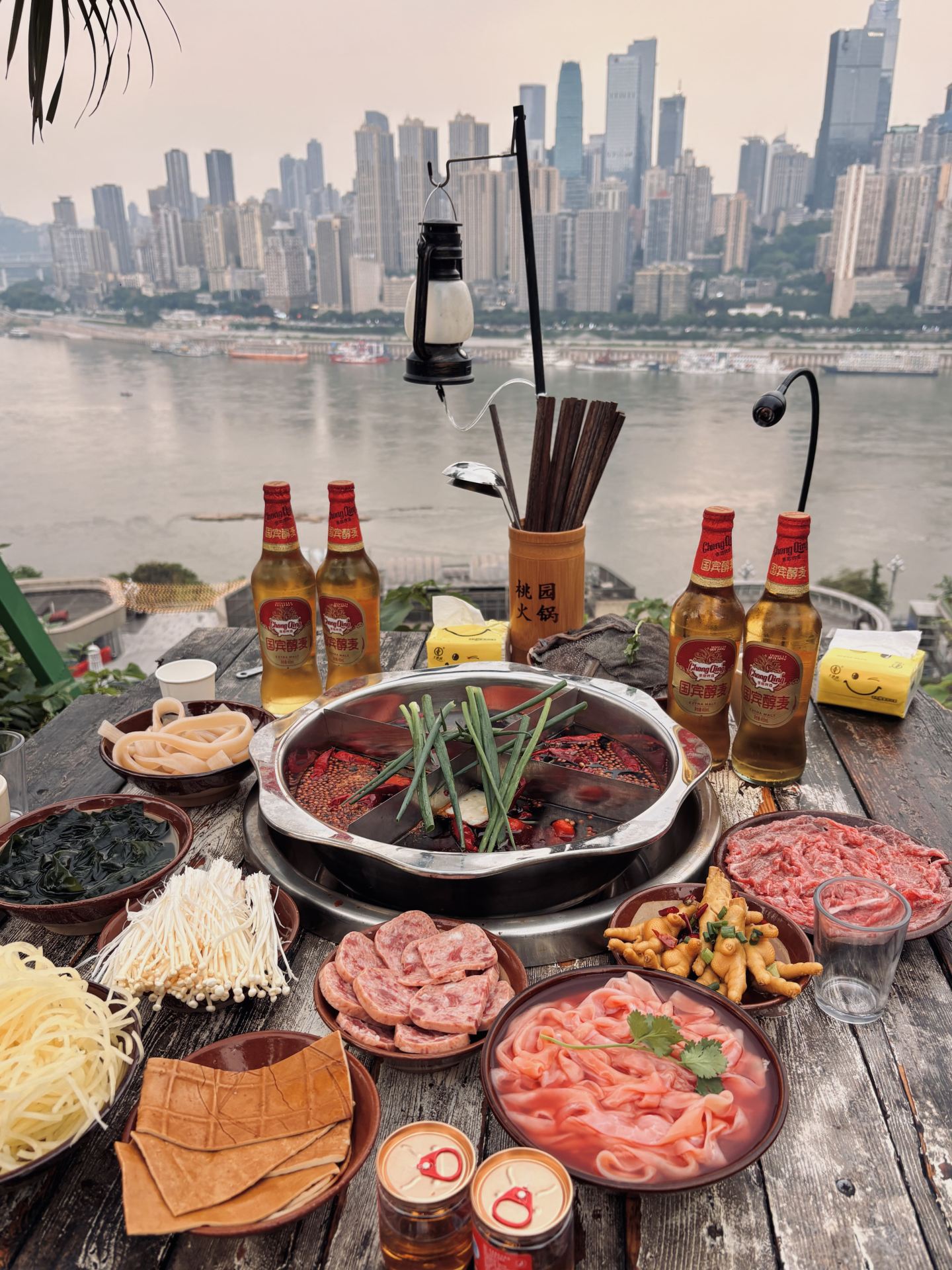 Demi menuntaskan keinginan ini, aku nemu Taoyuan Hotpot yang lokasinya di atas bukit.

Pengalaman pribadi: Pemandangan sungainya oke banget, city light pas malam hari cantik, dan pelayanannya lumayan oke. Rasa hotpot-nya sih standar aja, tapi poin plusnya harganya terjangkau banget. Ada live music juga mulai jam 7 malam. Buat kalian yang hobi foto-foto, tempat ini oke banget buat dapet spot foto yang estetik.

Bulan Mei hawanya agak gerah dan ada nyamuk, tapi angin sepoi-sepoi di atas bukit seger banget. Makan hotpot di sini bener-bener perjuangan buat kaki. Aku pribadi paling males naik tangga atau bukit, tapi ya namanya juga di Chongqing, isinya emang tanjakan semua, jadi ya udahlah ya gas aja.

Navigasi ke 📍Taoyuan Hotpot · View night scenery and listen to folk songs (di seberang Exit 3C Stasiun Light Rail Shanghao, Distrik Nan'an, Chongqing).

Nanti bakal kelihatan tangga dinding merah. Ikutin aja jalannya ke atas, terus bakal ada papan petunjuk warna-warni. Ikutin arah panah Taoyuan Hotpot dan lanjut naik tangga terus. Pas ketemu patung Totoro, terus aja naik ke atas. Tenang aja, banyak petunjuk jalannya kok. Begitu nyampe pintu masuk, stafnya bakal kasih segelas air buat ngelepas haus 🤣 Pengalaman yang unik dan kocak banget sih ini.