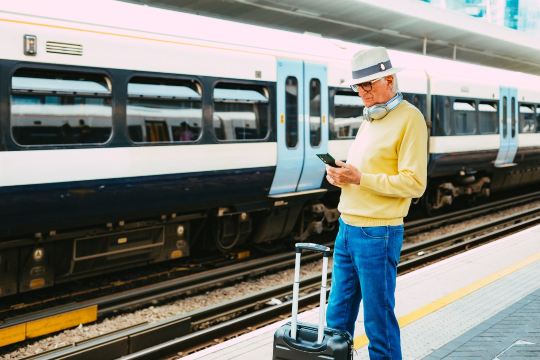 Senior traveler checking phone beside luggage at a train station platform.