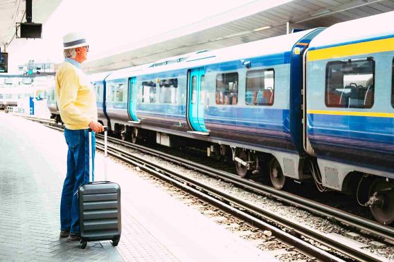 Elderly traveler with suitcase waiting beside blue train.