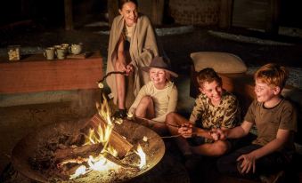 a group of people are sitting around a fire , with one person holding a stick at Paperbark Camp