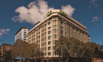 a large , modern building with a green sign on top is surrounded by trees and people at Vibe Hotel Sydney