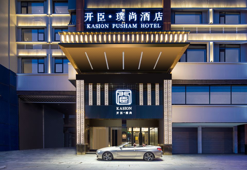 There is a hotel entrance with an illuminated sign above it, and there is another building in front of it at Kaison Pusham Hotel