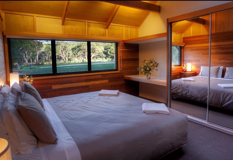 a modern bedroom with wooden walls , large windows offering views of the outdoors , and two beds made up with white linens at Girraween Environmental Lodge