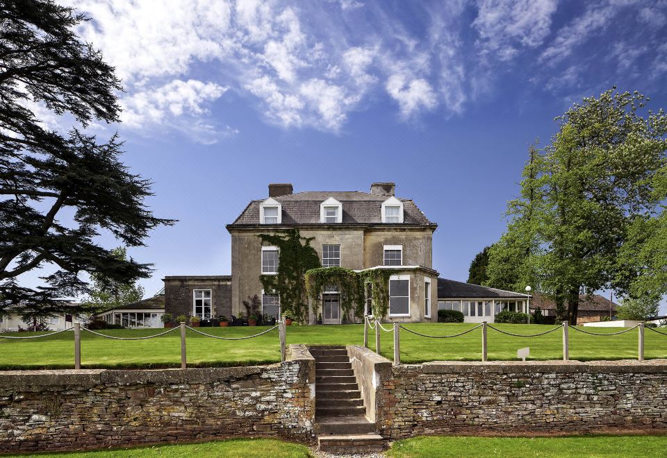 a large stone house surrounded by a grassy field , with trees in the background and stairs leading up to the front door at Mercure Bristol North the Grange Hotel