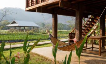 a wooden house with a hammock hanging from the porch , surrounded by greenery and a view of a farm in the distance at Lhongkhao Samoeng by Chi Villa