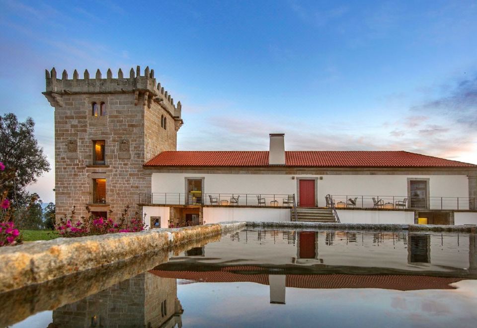a traditional stone building with a red roof , surrounded by a tranquil pond and a clear blue sky at Torre de Gomariz Wine & Spa Hotel