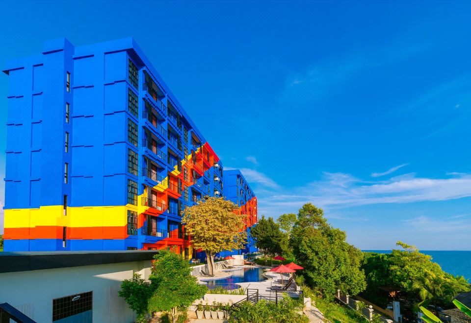 a large , multi - colored building with a blue and yellow facade is surrounded by trees and has a clear blue sky at Play Phala Beach Rayong