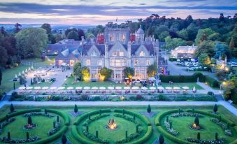 a large , ornate building with multiple windows and a domed roof , surrounded by lush greenery and illuminated at dusk at De Vere Tortworth Court