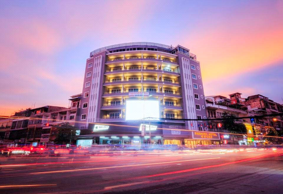 a city street at dusk , with a tall building in the background and several cars driving by at Paradise Hotel