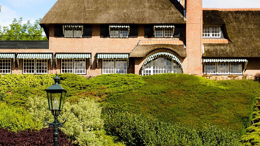 a brick house with a thatched roof and a porch , surrounded by bushes and greenery at Fletcher Hotel Restaurant de Wipselberg-Veluwe