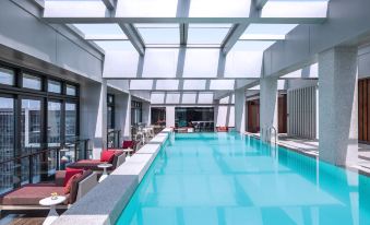 There is a spacious swimming pool surrounded by chairs and tables, located adjacent to an indoor lounge at Cordis Shanghai Hongqiao