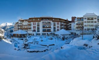 a large hotel surrounded by snow - covered trees , with several cars parked in the parking lot at Schlosshotel Fiss