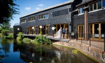 a wedding couple standing on a wooden bridge over a river , surrounded by lush greenery and modern black buildings at Tewinbury