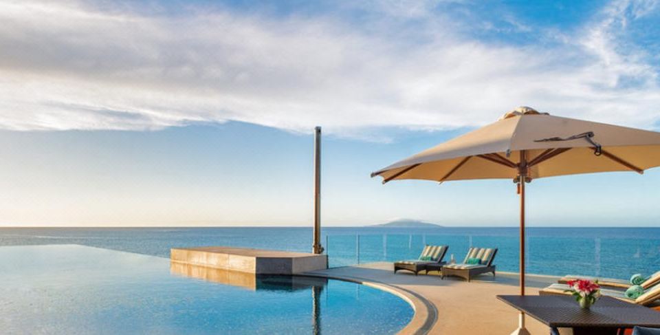 a large swimming pool with two lounge chairs and an umbrella overlooks the ocean and mountains at The Wakaya Club & Spa