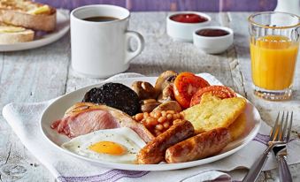 a plate of breakfast food , including bacon , eggs , beans , sausage , and toast , is placed on a table with a cup of coffee at Premier Inn Tewkesbury