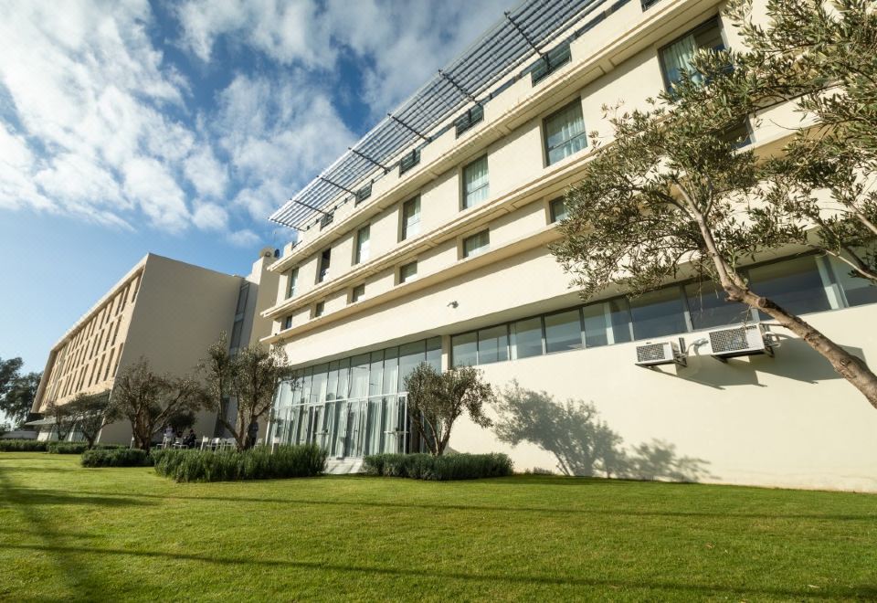 a large white building with a green lawn and trees in front of it , under a partly cloudy sky at Onomo Hotel Casablanca Airport