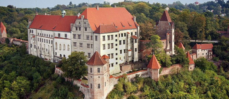 an old castle with a red roof , surrounded by trees and buildings , and a smaller castle tower at Achat Hotel Kaiserhof Landshut