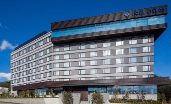 a modern hotel building with a large glass window , surrounded by trees and grass , under a clear blue sky at HOTEL MYSTAYS Fuji Onsen Resort