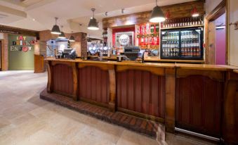 a bar with a wooden counter and shelves of bottles behind it , under pendant lights at Premier Inn Tewkesbury