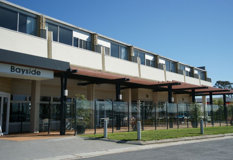 a building with a large glass entrance and black shutters , surrounded by trees and a clear sky at Bayside Hotel