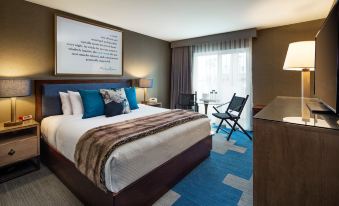 a large bed with blue and brown bedding is in a room with a window , chair , and framed poster at Hotel Hermosa