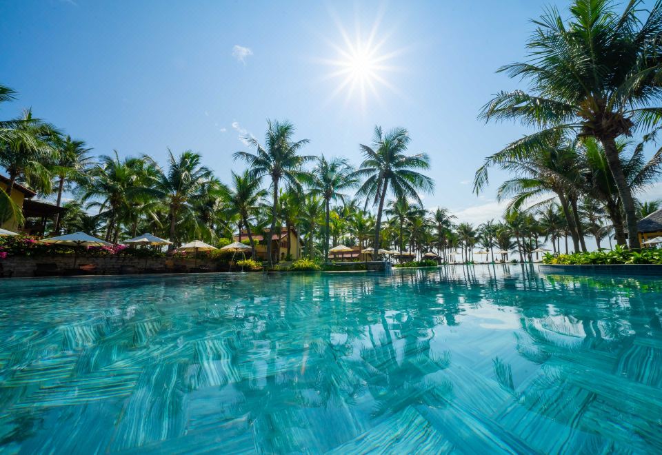 a large outdoor swimming pool surrounded by palm trees , with the sun shining brightly in the sky at Pandanus Resort