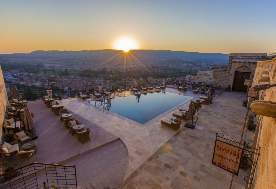 a large outdoor pool is surrounded by lounge chairs and a stone building with a sunset view at Kayakapi Premium Caves Cappadocia