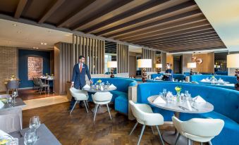 a modern dining room with blue and white furniture , including tables , chairs , and couches , as well as a waiter standing nearby at Clayton Hotel Chiswick
