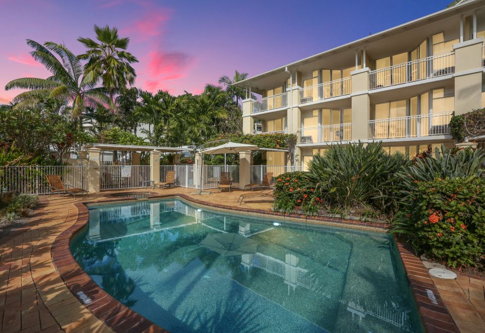 a large swimming pool is surrounded by a brick patio and palm trees , with a building in the background at On the Beach Apartments