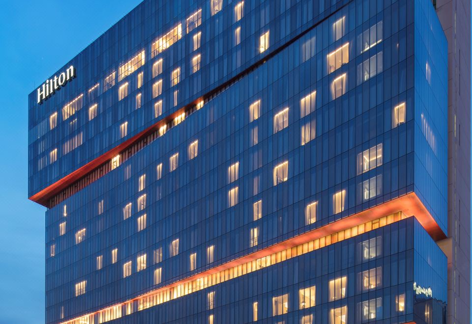 At night, a large illuminated building with multiple windows and a clock is visible from the outside at Hilton Guangzhou Tianhe