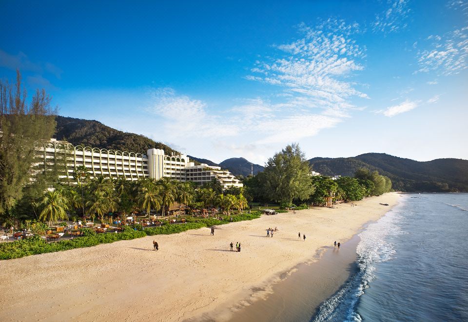 a beach with people walking and a large hotel in the background , surrounded by mountains at PARKROYAL Penang Resort