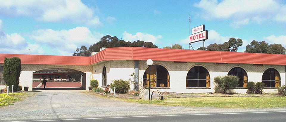 a motel with a red roof , surrounded by trees and a clear sky , and a sign above the entrance at Charlton Motel