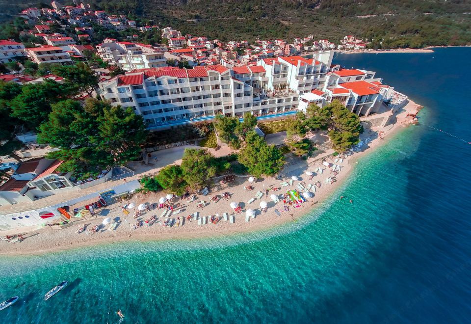 a beach resort with a large white building surrounded by clear blue water and white sand , with numerous people enjoying their time on the beach at Tui Blue Makarska