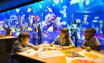 Children are seated at a table with their teacher, while other children in the background observe an exhibit at The Parisian Macao
