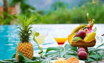 a table with a pineapple , fruit , and a cocktail in front of a pool surrounded by greenery at Tam Coc La Montagne Resort & Spa